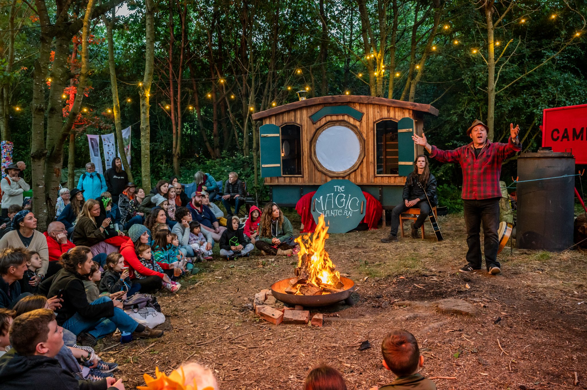 A large group of people are gathered on one side of a campfire listening to Ian Douglas perform fables.