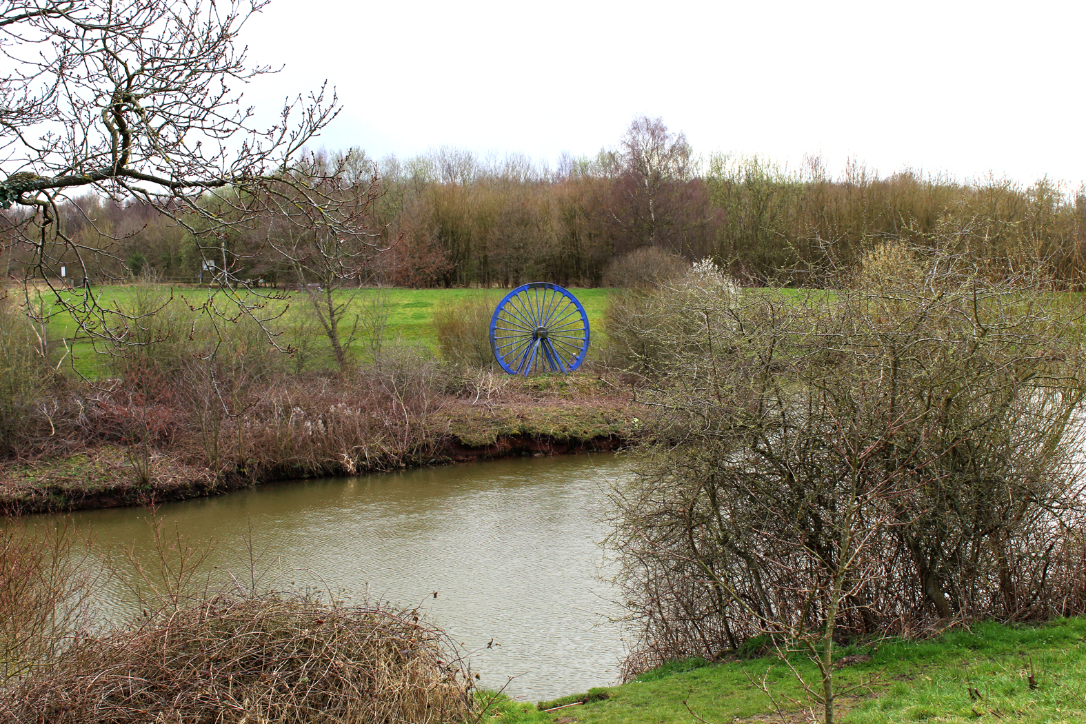 A river between two fields with bare trees and bushes either side. A blue model of a mining wheel is on the far bank.