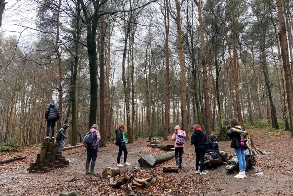 A group of people stand in a woodland clearing. There are wooden sculptures on the ground around them.