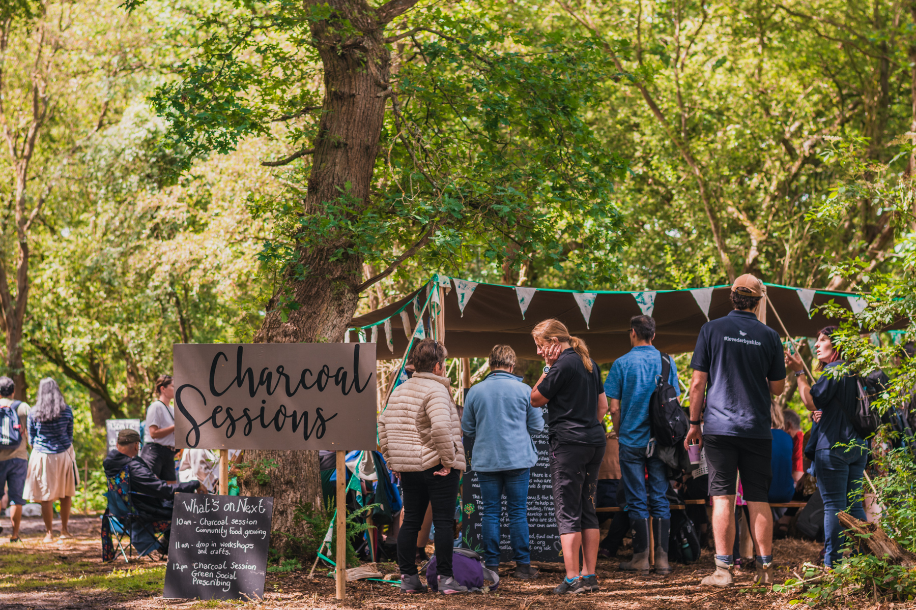 People gathered under a shelter with a sign saying "Charcoal Sessions" in script in front of it.