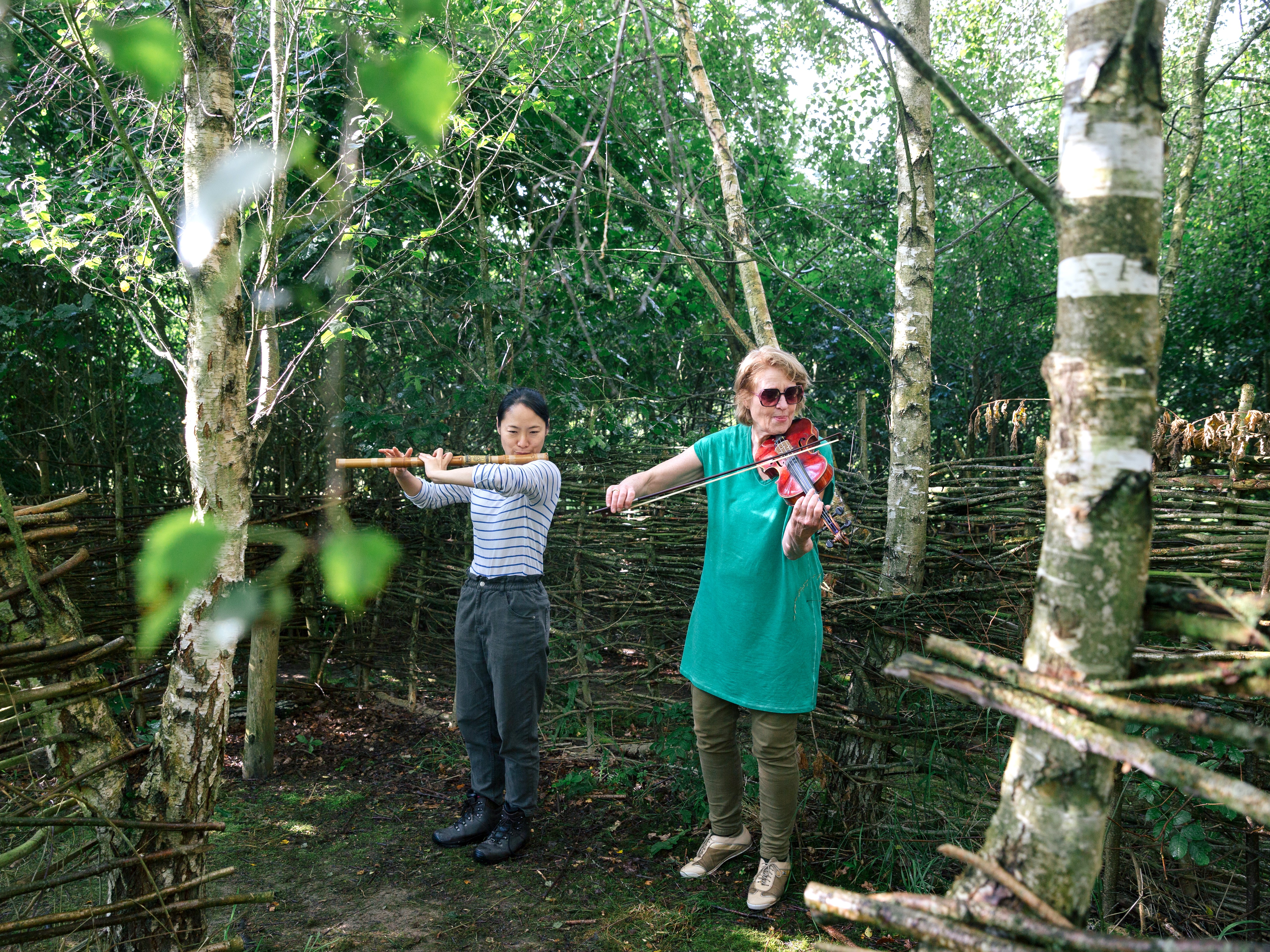 Two people playing a wooden flute and a violin in a woodland setting.