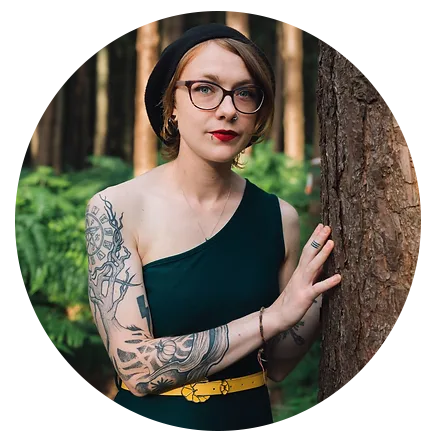 Sophie Sparham stands in a woodland touching a tree and looking at the camera, She is wearing bright red lipstick and a black beret.