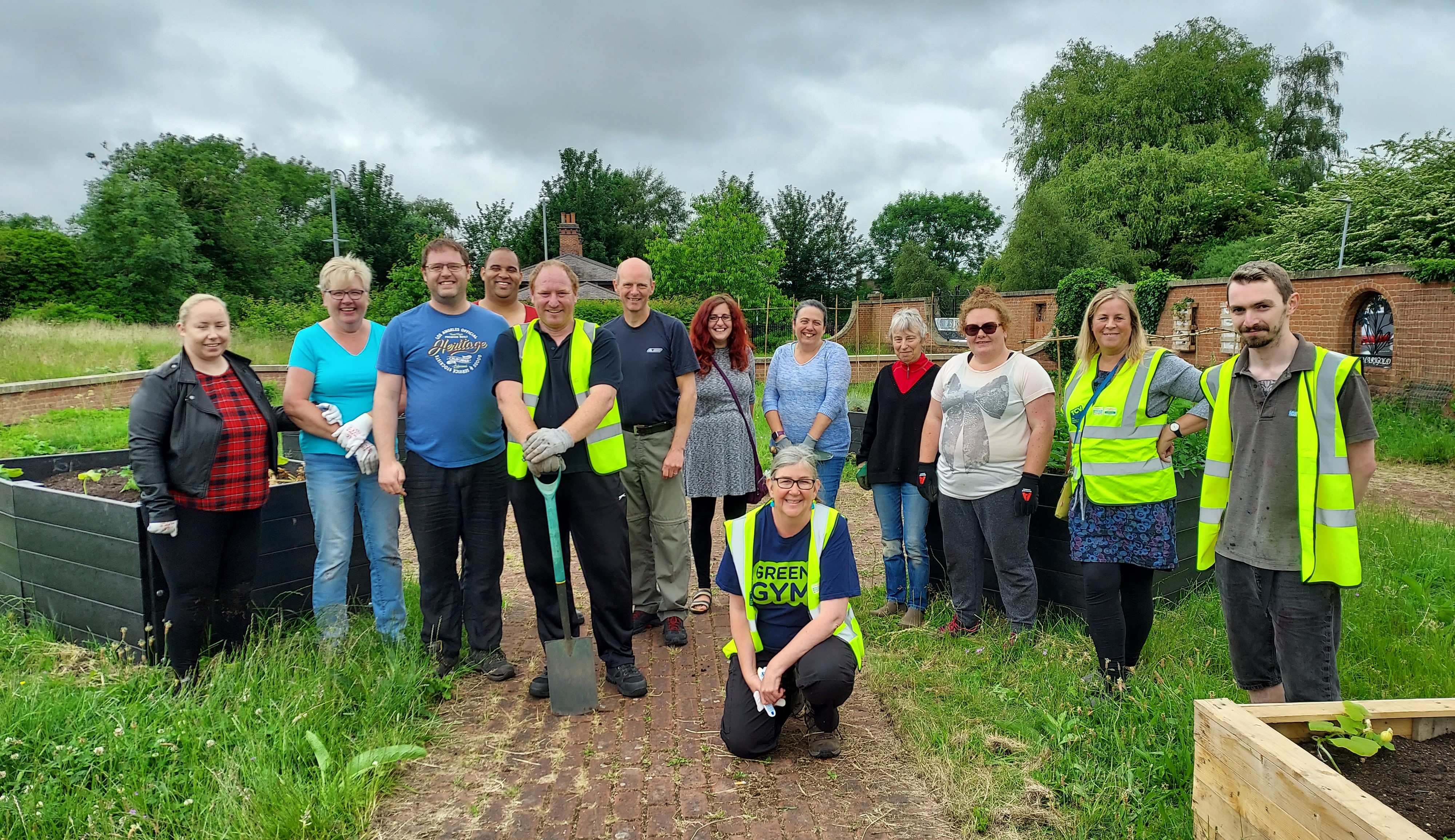 A group of people smiling at the camera in a garden with raised beds around. Some are wearing hi-vis jackets and holding spades.