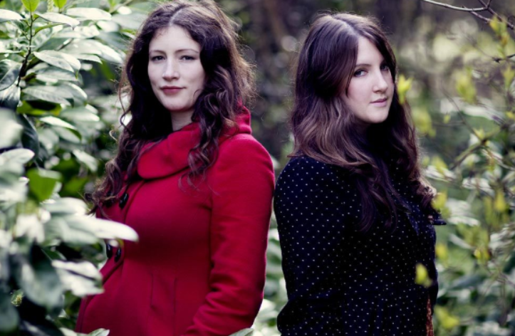 The Unthanks: two women with long brown hair looking at the camera whilst stood between trees.