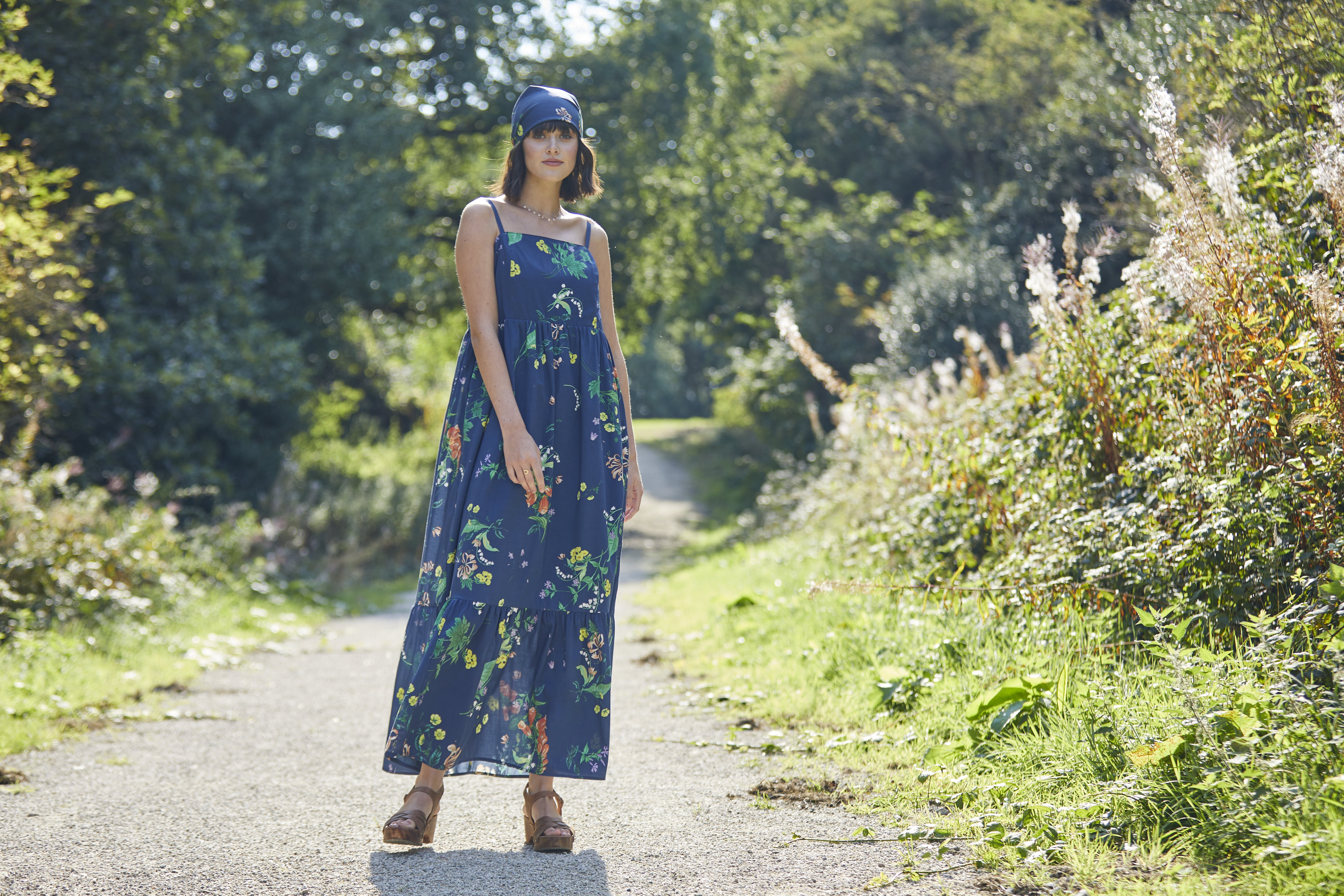 A woman in a long, floaty blue dress with flowers on stands in the middle of a path. She has a matching bandana on her head. 