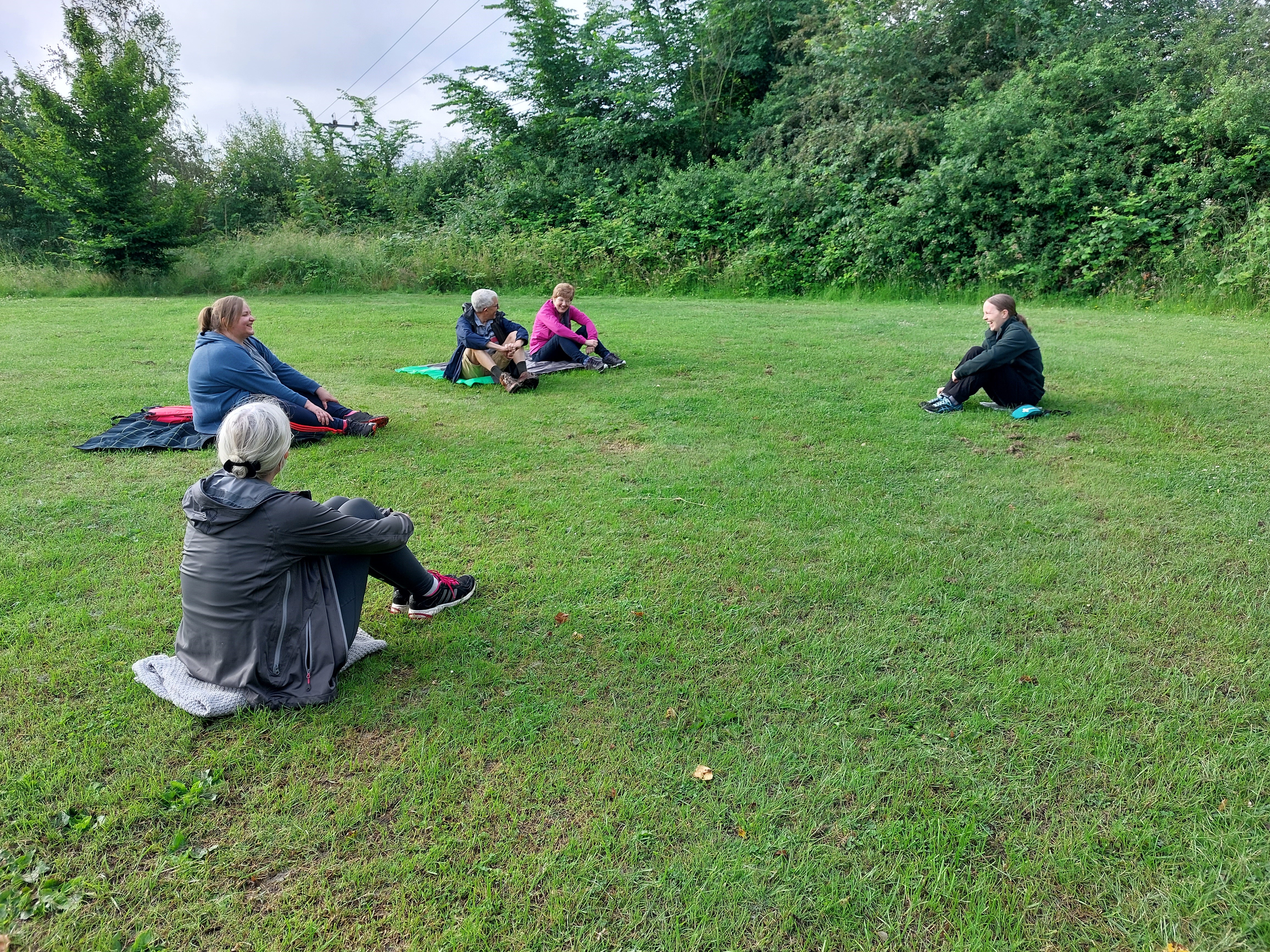 People sitting on towels and yoga mats in a field with trees around the edge